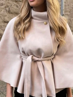 Beige kappa med knytband - Stilren beige kappa med hög krage och elegant knytband i midjan. Kappan har vida ärmar och en dekorativ knapp vid halsen. Perfekt för dig som gillar minimalistisk och chic stil.