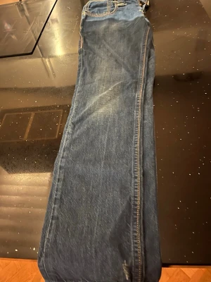 Levi's 501 blå jeans med röd patch - Klassiska blå Levi's 501 jeans med rak passform och tydlig röd Levi's patch bak. Jeansen har fem fickor, kontrastsömmar och är tillverkade i slitstarkt bomullsjeansmaterial. Perfekta för en avslappnad och tidlös look.