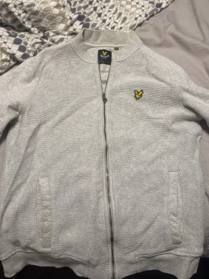 Ljusgrå ziptröja från Lyle & Scott - Säljer en ljusgrå ziptröja från Lyle & Scott med gul logga på bröstet. Tröjan har ribbade muddar, ståkrage och två fickor framtill. Perfekt för en avslappnad och stilren look.