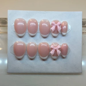 Rosa och vita lösnaglar med rosett - Snygga lösnaglar i en mjuk rosa nyans med vita tippar. Två av naglarna har en söt rosa rosett som detalj. Perfekt för en elegant och lekfull look.