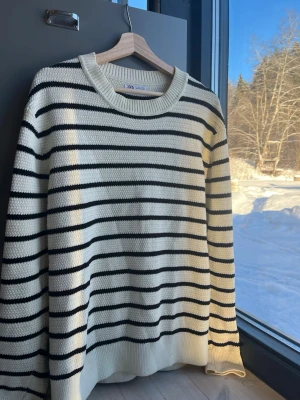 Randig stickad tröja från Zara strl L - Snygg stickad tröja från Zara i vitt med svarta horisontella ränder. Tröjan har rund halsringning och långa ärmar. Perfekt för lager-på-lager och enkel att matcha med jeans eller kjol.