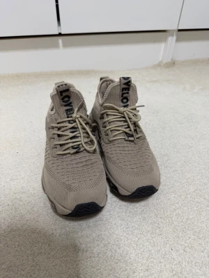 Beige sneakers med stickad ovandel - Snygga beige sneakers med stickad ovandel och cool textdetalj på plösen. Skorna har snörning och en sportig sula med transparenta partier. Perfekta för dig som gillar en clean och modern stil.