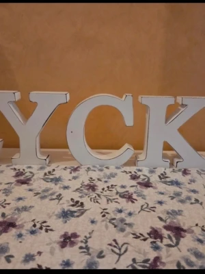 Vita träbokstäver LYCKA - Dekorativa bokstäver i vitt trä som tillsammans bildar ordet 'LYCKA'. Bokstäverna har en lätt sliten look för en charmig vintagekänsla och passar perfekt som inredningsdetalj på hylla eller bord.