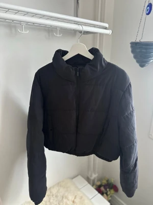 Svart croppad pufferjacka - Snygg svart croppad pufferjacka med hög krage och dragkedja framtill. Jackan har quiltad design och en loose pass sopm ger en avslappnad vibe. Perfekt för kalla dagar när du vill vara både varm och trendig.