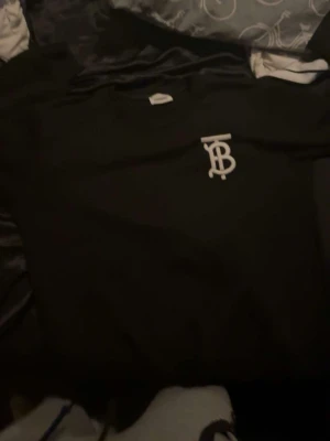 Svart sweatshirt från Burberry - Stilren svart sweatshirt från Burberry med vitt TB-monogram broderat på bröstet. Klassisk rund halsringning och avslappnad passform. Perfekt för dig som gillar exklusiva streetwear-plagg.