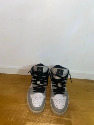 Nike Air Jordan 1 Mid vit/svart/grå - Snygga Nike Air Jordan 1 Mid sneakers i vitt, svart och grått. Klassisk siluett med svart swoosh, svarta skosnören och perforerad tå. Tillverkade i skinn med ikoniska Jordan Wings-loggan på sidan. Perfekta för dig som gillar streetwear och tidlös stil.