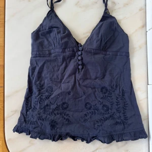 Mörkblå broderat linne med sidenrosett i ryggen - Supersöt mörkblå linnetopp med smala axelband, broderade blommor nertill och volangkant. Toppen har dekorativa knappar framtill och knytning i ryggen för extra snygg passform. Perfekt för sommarens alla tillfällen!