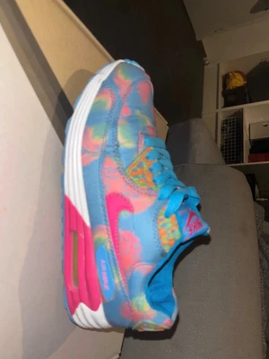 Nike Air Max i blått och rosa mönster - Färgglada Nike Air Max sneakers med blå bas och rosa, gula samt gröna detaljer i ett lekfullt mönster. Skon har vit och rosa sula med synlig Air Max-dämpning, snörning och rund tå. Perfekt för dig som vill sticka ut med dina sneakers.