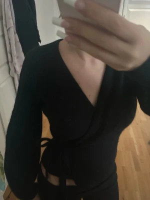 Svart omlottkofta från & other stories  - Trendig svart omlottkofta från & other stories  med långa ärmar och knytband i midjan. Koftan har en djup v-ringning och puffiga axlar som ger en snygg siluett. Perfekt att styla över ett linne eller topp för en cool look.