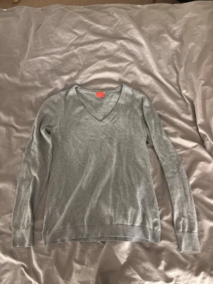 Ljusgrå stickad V-ringad tröja - En stilren ljusgrå stickad tröja med V-ringning och långa ärmar. Tröjan har ribbade muddar vid ärmslut och nederkant, vilket ger en snygg passform. Perfekt att matcha med jeans eller kjol.