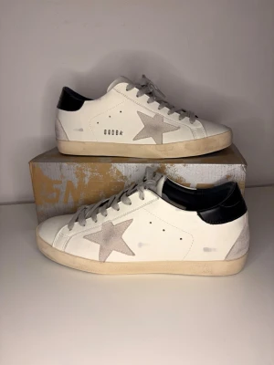 Golden Goose Superstar + box & dustbag - Säljer ett par Golden Goose Superstar i klassisk vit färg med grå stjärna och svart häl. Skorna är endast testade inomhus, i mycket fint skick.  ✔️ Original låda ingår ✔️ Original dustbag ingår ✔️ Ikonisk Superstar-modell ✔️ Stilrena och lätta att matcha  Perfekt för dig som vill ha premium sneakers med exklusiv känsla.