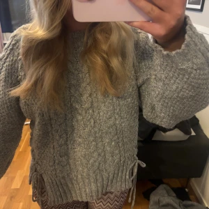 Grå kabelstickad oversized tröja - Mysig grå stickad tröja med kabelstickat mönster framtill och grov struktur på ärmarna. Tröjan är oversized med långa ärmar och har snörningsdetaljer i sidorna. Perfekt för kalla dagar och enkel att styla till olika outfits.