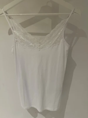 Vit spetslinnetopp från Vila storlek S - Säljer en vit linnetopp från Vila med tunna axelband och vacker spetsdetalj längs urringningen. Toppen har en feminin v-ringad form och är perfekt att styla lager på sopmaren. Materialet känns mjukt och lätt mot huden.