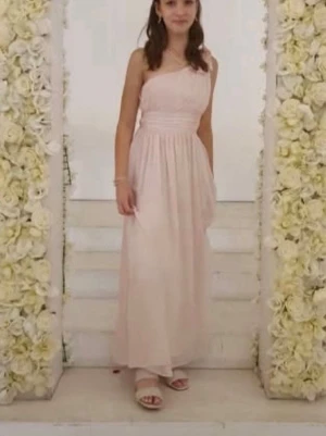 Ljusrosa långklänning med oneshoulder - Säljer en elegant ljusrosa långklänning med oneshoulder och fin blomdetalj på axeln. Klänningen har ett plisserat liv och faller mjukt ner mot golvet. Perfekt för speciella tillfällen när du vill känna dig extra fin.