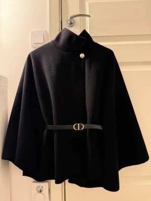 Svart cape-kappa från Chelsea - Stilren svart cape-kappa från Chelsea med hög krage och elegant bälte i midjan med gulddetalj. Kappan har en knapp framtill och ett svepande, löst fall som ger en cool siluett. Perfekt för dig som vill ha en trendig look.