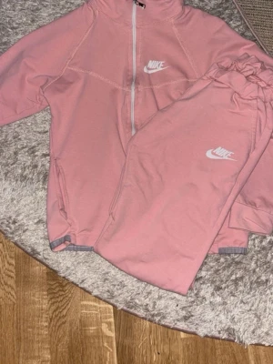Rosa mjukisbyxor från Nike - Säljer ett par snygga rosa mjukisbyxor från Nike med klassisk logga på benet. Byxorna har resår i midjan och är gjorda i mjukt material som passar perfekt för chill eller träning. Matchande hoodie med dragkedja och huva ingår.