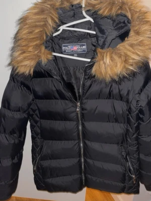 Svart dunjacka Polo Club pälsdetalj - Snygg svart dunjacka från Polo Club Saint Vincent med quiltad design och huva med fejkpäls. Jackan har dragkedja framtill och detaljer på ärmen. Perfekt för kalla dagar och ger en cool look.