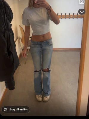 Ljusblå bootcut jeans med hål - Säljer ett par ljusblå bootcut jeans från Nelly med stora hål över knäna. Jeansen har låg midja och klassisk femficksdesign. Perfekta för en avslappnad och trendig look.
