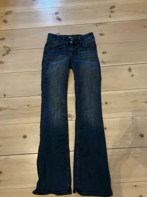 Mörkblå bootcut jeans med låg midja - Snygga mörkblå jeans med bootcut och låg midja. Klassisk femficksmodell med knappar och dragkedja framtill samt fickor med lock bak. Perfekta för en avslappnad och trendig look. Lite skavda längst ner vid foten, ngt långa för mig som är 164 cm. 