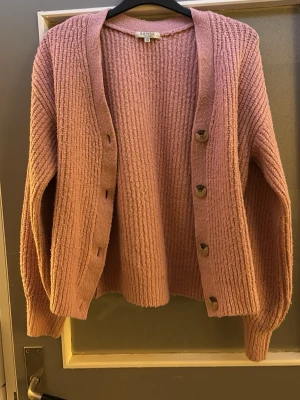 Rosa ribbstickad kofta  - Mysig rosa kofta med ribbstickad struktur och stora knappar framtill. Koftan har en avslappnad passform, långa ärmar och breda muddar. Perfekt för lager-på-lager och enkel att styla till olika outfits. Storlek 146/152 S