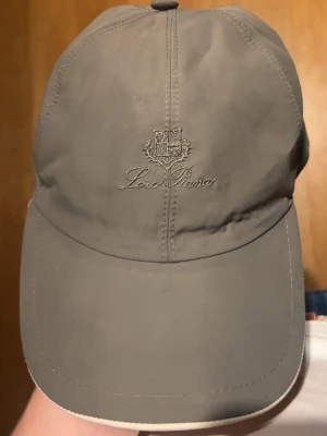 Grön keps från Loro Piana - Snygg grön keps från Loro Piana med broderad logga framtill och klassisk böjd skärm. Insidan har ett rutigt foder och etikett med 'Storm System'. Perfekt accessoar för en stilren look.