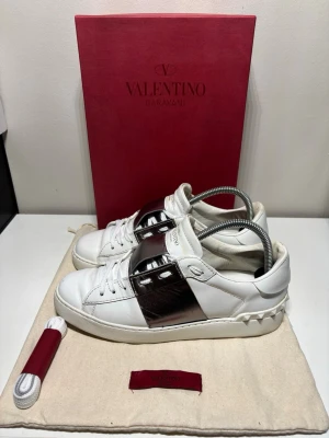 Valentino Garavani Open sneakers vit/silver - Snygga Valentino Garavani Open sneakers i vitt skinn med en bred metallic silverrem över mitten. Klassisk låg modell med rund tå, snörning och ikoniska nitar på hälen. Kommer med extra skosnören och originalförpackning. Storlek 38,5 och i bra skick, box, dustbag och snören ingår