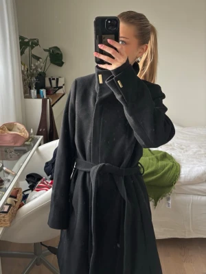 Svart kappa med  krage - Stilren svart kappa med hög krage och snygga guld-detaljer vid halsen. Kappan har ett knytbälte i midjan och långa ärmar. Perfekt för kyligare dagar och enkel att matcha med olika outfits.