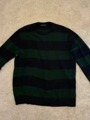 Randig stickad tröja i grönt och svart - Snygg stickad tröja med breda horisontella ränder i mörkgrönt och svart. Tröjan har rund halsringning, långa ärmar och ribbade muddar vid ärmslut och nederkant. Perfekt för dig som gillar klassisk men ändå cool stil.