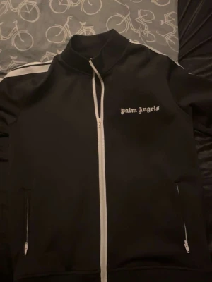 Svart track jacket från Palm Angels - Snygg svart track jacket från Palm Angels med vit dragkedja och vita detaljer på ärmarna. Jackan har hög krage, två fickor med dragkedja och logga tryckt på bröstet. Perfekt för en sportig och trendig look.