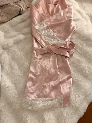 Ljusrosa sidenmorgonrock med spets - Supersöt ljusrosa morgonrock i glansigt sidenmaterial med vita spetsdetaljer vid ärmslut och nederkant. Knytband i midjan för en snygg passform. Perfekt för mysiga hemmakvällar eller när du vill känna dig extra lyxig.