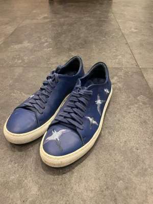 Blå sneakers från Axel Arigato - Snygga blå sneakers från Axel Arigato med broderade fåglar på sidan. Skorna har rund tå, snörning och vit platt sula. Tillverkade i skinn och har en stilren design som sticker ut med de broderade detaljerna.