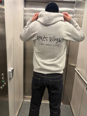 Grå hoodie Après Royale - Snygg grå hoodie från Après Royale med svart tryck på ryggen där det står 'Après Royale' och 'Nølt Nordic Alpine'. Hoodien har huva och en avslappnad passform, perfekt för en chill stil.