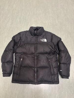 Svart pufferjacka från The North Face - Säljer en svart pufferjacka från The North Face med hög krage och dragkedja framtill. Jackan har två snedställda fickor med dragkedja och logga broderad på bröstet. Perfekt för kalla dagar och riktigt snygg streetstil.
