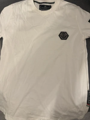 Vit t-shirt från Philipp Plein - Vit t-shirt från Philipp Plein med svart logomärke på bröstet och ett svart märke med silvertext på ärmen. Klassisk rund halsringning och stilren design. Perfekt för dig som gillar exklusiva streetwear-plagg.