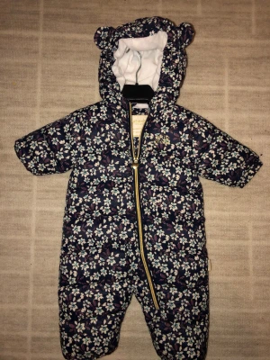 Blommig pufferjacka från Petite Chérie - Supersöt blommig pufferjacka från Petite Chérie med huva och små öron på toppen. Jackan har ett quiltat mönster, dragkedja i guld och är perfekt för kyliga dagar. Mörkblå botten med vita och lila blommor ger en gullig look.