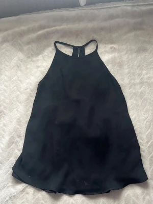 Svart blus med rosett - Stilren svart blus i ett lätt, flödande tyg med hög halsringning och öppen rygg. Blusen är ärmlös och har en dekorativ rosett baktill som ger en extra snygg detalj. Perfekt att matcha med jeans eller kjol för en trendig look.