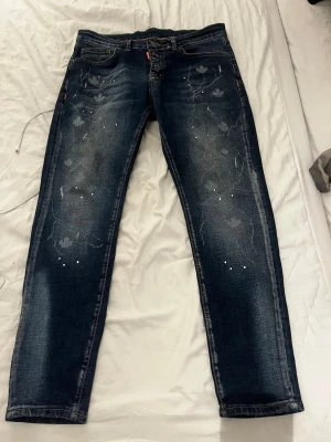 Mörkblå Dsquared2 jeans med tryck - Snygga mörkblå jeans från Dsquared2 med unikt löv- och stänkmönster framtill. Jeansen har raka ben, klassisk femficksdesign och röda detaljer vid gylfen. Perfekta för dig som vill sticka ut med en cool streetstil.