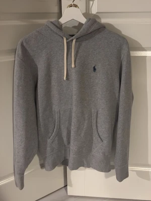 Grå hoodie från Polo Ralph Lauren - Klassisk grå hoodie från Polo Ralph Lauren med vit dragsko i huvan och den ikoniska blå loggan broderad på bröstet. Hoodien har känguruficka framtill och är tillverkad i mjukt bomullsmaterial. Perfekt för en avslappnad och stilren look. Skriv för bild på! 