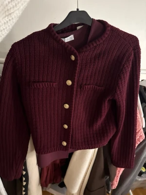 Vinröd ribbstickad kofta med guldknappar - Vinröd ribbstickad kofta med rund hals och dekorativa guldknappar framtill. Kortare modell med långa ärmar och två fejkfickor på bröstet. Perfekt för dig som vill ha en stilren och trendig look.