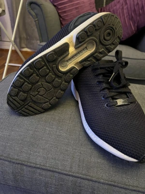 Svarta Adidas sneakers med mesh - Svarta Adidas sneakers med ovandel i mesh och snörning. Skorna har en vit sula med svart yttersula och diskret Adidas-logga på plösen. Perfekta för en sportig och avslappnad stil.