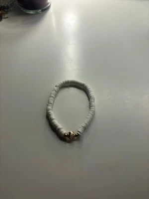 Armband - Snyggt armband med vita platta pärlor och en guldfärgad snäcka i mitten. Perfekt accessoar för en fräsch och somrig look. Elastiskt band gör det enkelt att ta på och av.