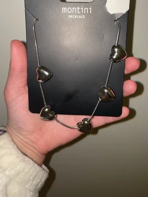 Halsband med hjärtan från Montini - Snyggt halsband från Montini med fem stora, silverfärgade hjärtan på en tunn kedja. Hjärtana har en blank finish och halsbandet ger en trendig och lekfull vibe. Perfekt accessoar för att lyfta din outfit med en cool detalj.
