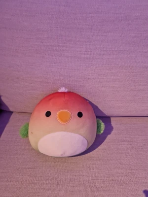 Squishmallows fågel mjukisdjur liten - Supermjuk och rund fågel från Squishmallows med persikofärgad ovansida, vit mage, orange näbb och gröna vingar. Liten vit tofs på huvudet och broderade ögon. Perfekt att krama eller ha som dekoration i soffan.