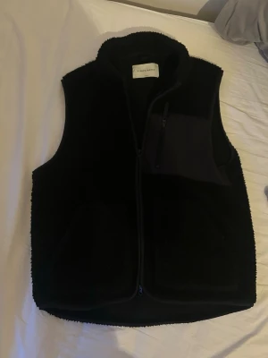 Svart teddyväst från A Day's March - Svart teddyväst från A Day's March med hög krage och dragkedja framtill. Västen har två stora fickor och en bröstficka i kontrasterande tyg med dragkedja. Perfekt för lager på lager och ger en avslappnad streetwear-vibe.