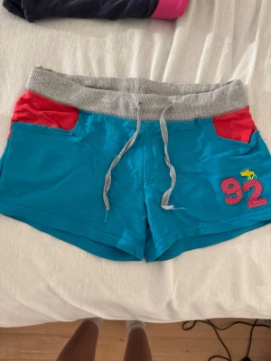 Vintage blåa lowwaist shorts - Säljer ett par blå shorts med grå midja och snörning, röda fickor och broderat tryck med siffran 92 och en gul älg på vänster ben. Perfekta för chill dagar eller träning. Mjukt material och relaxed fit.