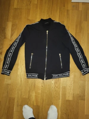 Svart tröja från Balmain Paris - Orginal svart tröja från Balmain Paris med vita logotexter längs ärmar och nederkant. Jackan har silvriga dragkedjor framtill och på fickorna samt ribbade muddar. Snygg och stilren design som sticker ut med tydliga Balmain-detaljer.