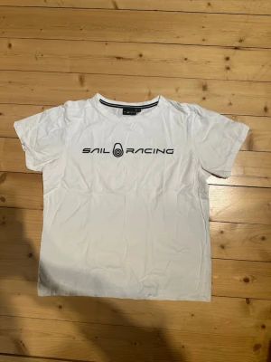 Vit Sail Racing t-shirt med tryck - Vit t-shirt från Sail Racing med svart logotyptryck på bröstet och en liten logga i nacken. Klassisk rund hals och korta ärmar. Tillverkad i mjuk bomull för en skön känsla. Perfekt till jeans eller shorts. Tveka inte att skriva  vid frågor eller funderingar 