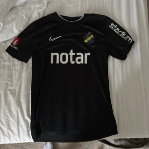 Svart AIK tröja Nike - AIK:s officiella svarta matchtröja från Nike med klubbmärke på bröstet, vita tryck med sponsorloggor och detaljer. Tröjan är i lätt och ventilerande material, perfekt för fotbollsträning eller match. Klassisk rund hals och korta ärmar, tröjan är andvänd av rashidi och fick den av han 2020