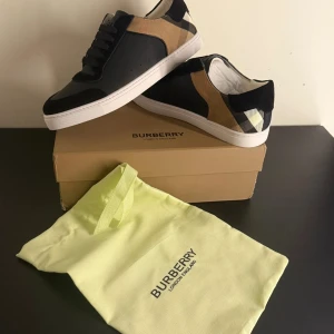 Burberry sneakers med rutigt mönster - Snygga Burberry sneakers med klassiskt rutigt mönster i beige, svart och grått. Skorna har en rund tå och snörning framtill. Perfekta för en stilren look.