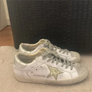  sneakers med glitterstjärna - Snygga vita sneakers  med grå snörning och silverdetaljer på hälen. På sidan finns en stor glittrig stjärna i guld som ger extra attityd. Skorna är låga och har rund tå samt platt sula. Perfekta för dig som gillar en unik och trendig look.
